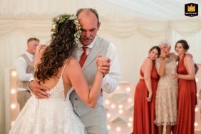 During a private marquee wedding in Dorset, the father and daughter share a dance at the center while, in the background, the bride’s teary-eyed mother is comforted by bridesmaids, capturing both joyful and emotional reactions within the same scene.